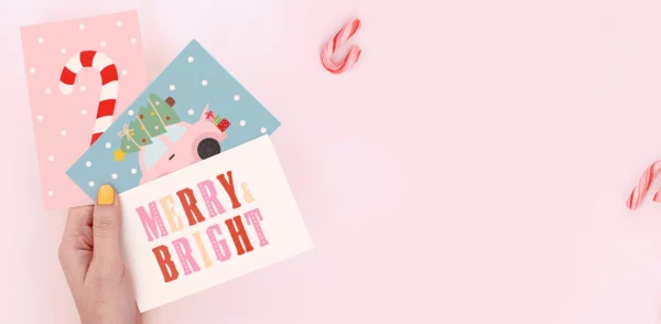 Hand som håller färgglada födelsedagskort med julmotiv och texterna ”Merry Bright” och ”Ho Ho Ho” på en julklapp.