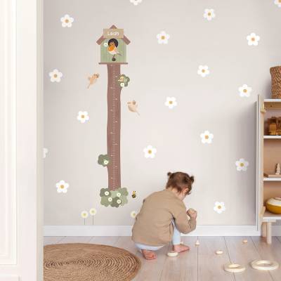En liten flicka leker på ett barnrum med en väggdekal i form av en fågelholk och en mätsticka, omgiven av blommiga dekorationer. Perfekt som väggdekor för barnerum.