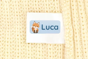 Personlig navnelapp med rävillustration och namnet Luca mot en ljus stickad bakgrund. Perfekt för märkning av barnkläder.
