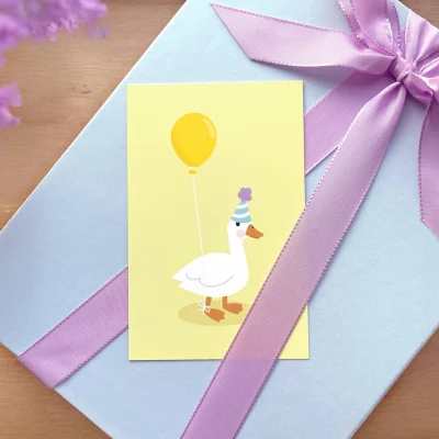 Fargerikt bursdagskort med en tegning av en glad ankh med ballong og festhatt, plassert på en lyseblå gaveboks med lilla bånd. Perfekt til barnebursdager.