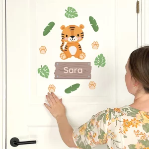 En kvinne henger opp en personlig dørdekorasjon med en søt tiger og navnet 'Sara'. Dekorasjonen er omkranset av grønne blader og poter, og passer perfekt til barnerommet. Oppdag vårt utvalg av veggdekor for barn og personlige navnelapper.