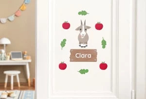 Dørdekor for barn med navnet Clara, illustrert med søte detaljer som en geit og røde epler, perfekt for barnerommet. Denne dekorasjonen gir et personlig preg til barnets rom og skaper en koselig atmosfære.