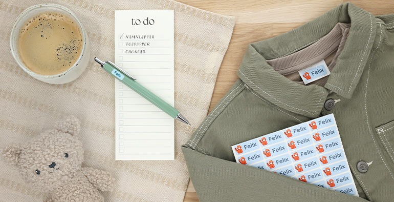 Navnelapper med navnet 'Felix' plassert på en bordflate sammen med en god kopp kaffe, en notatblokk med to-do-liste, en grønn penn, trådløse hodetelefoner, en kosedyr-bjørn, og et klesplagg. Perfekt for organisering og personlig tilpasning.