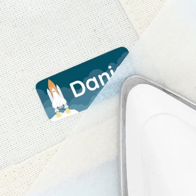 Stryklapp med navn og rakettmotiv, designet for barn, som viser hvordan personlig tilpasning kan brukes til klær og eiendeler. Dette produktet bidrar til å gjøre barnas ting unike og lette å identifisere.