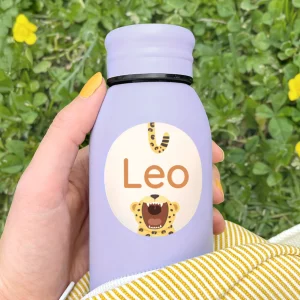 Person som holder en personlig navnelapp med navnet 'Leo' på en lila drikkeflaske, perfekt for barn. Navnelappen har en lekende løve-design, ideell for skoleutstyr og tilpassede produkter for barnerommet.