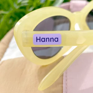 Minilapp med navnet 'Hanna' festet på gule solbriller, ideelt for barn. Perfekt for å personliggjøre eiendeler og unngå havari. Besøk vårt nettsted for å se flere navnelapper og barneprodukter.