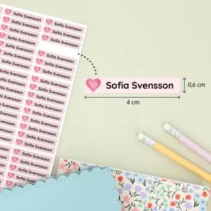 Navnelapper med navnet Sofia Svensson, designet med hjerte og rosa bakgrunn, vist på et ark. Lappene måler 4 cm x 0,6 cm og er perfekt for å merke ting til barn. To fargepennene og et mønstrete notatblokk er synlige i bakgrunnen, noe som gir en koselig og barnslig atmosfære. Ideelt for barnerom, bursdagsfeiringer og personlige gaver.