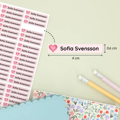 Navnelapper med navnet Sofia Svensson, designet med hjerte og rosa bakgrunn, vist på et ark. Lappene måler 4 cm x 0,6 cm og er perfekt for å merke ting til barn. To fargepennene og et mønstrete notatblokk er synlige i bakgrunnen, noe som gir en koselig og barnslig atmosfære. Ideelt for barnerom, bursdagsfeiringer og personlige gaver.
