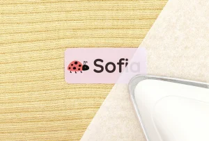 Navnelapp med myggdesign och namnet Sofia, placerad på en gul stickad textil, tillsammans med ett strykjärn. Perfekt för att märka kläder och tillhörigheter för barn.