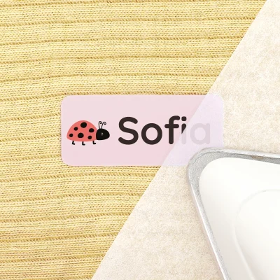Navnelapp med myggdesign och namnet Sofia, placerad på en gul stickad textil, tillsammans med ett strykjärn. Perfekt för att märka kläder och tillhörigheter för barn.