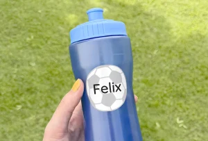 En hand håller en blå sportflaska med en personlig navnelapp, designad med en fotboll och namnet Felix. Bakgrunden är grön, vilket skapar en utomhuskänsla. Perfekt för aktiva barn som spelar fotboll och behöver hålla sig hydrerade.