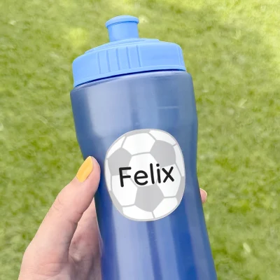 En hand håller en blå sportflaska med en personlig navnelapp, designad med en fotboll och namnet Felix. Bakgrunden är grön, vilket skapar en utomhuskänsla. Perfekt för aktiva barn som spelar fotboll och behöver hålla sig hydrerade.