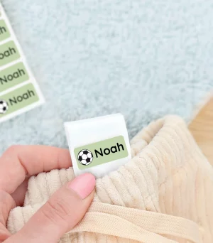 Namnlappar med fotbollsmotiv som sticker ut från en klädesdetalj, med temat för barnprodukter. I bakgrunden syns en grön barnskor. Perfekt för att personifiera barnens kläder och hitta dem lättare.