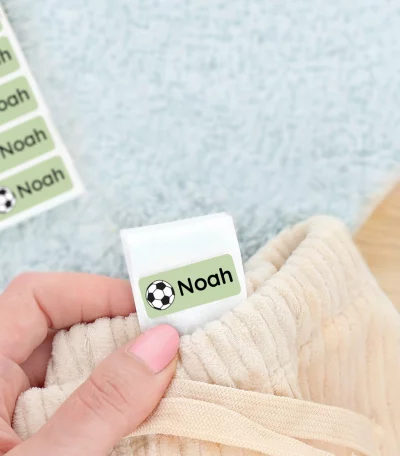 Namnlappar med fotbollsmotiv som sticker ut från en klädesdetalj, med temat för barnprodukter. I bakgrunden syns en grön barnskor. Perfekt för att personifiera barnens kläder och hitta dem lättare.
