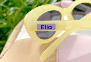 Gula solglasögon med personligt namnklistermärke, perfekt för barn. Omgiven av gröna gräs och mjuk textil, idealisk för sommarens lek och utflykter.
