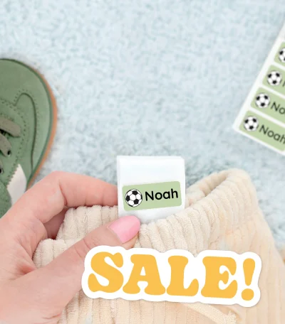 Färgglada namnlappar för barnprodukter mot en mjuk bakgrund. Bilden visar en hand som håller i en namnlapp med texten Noah designad för att fästa på kläder eller skor. Bakom syns barnskor och en lapp med fler namnlappar. Produkten passar perfekt för att organisera barnens tillhörigheter och håller dem unika.