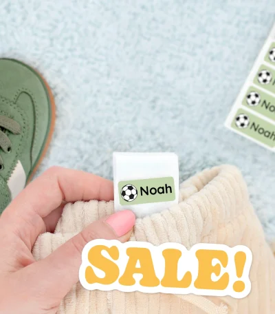 Färgglada namnlappar för barnprodukter mot en mjuk bakgrund. Bilden visar en hand som håller i en namnlapp med texten Noah designad för att fästa på kläder eller skor. Bakom syns barnskor och en lapp med fler namnlappar. Produkten passar perfekt för att organisera barnens tillhörigheter och håller dem unika.