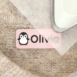 Färggrann navnlabel med en söt pingvin och namnet Olivia, som ligger på en mjuk yta. Perfekt för att personifiera barnets tillhörigheter och ge en lekfull touch. Idealisk för att användas på kläder, väskor och andra föremål.