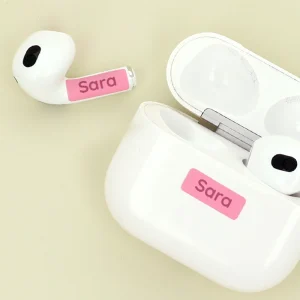Vita trådlösa hörlurar med personliga namnlappar i rosa, som visar namnet Sara, ligger bredvid ett öppet etui. Perfekt för att hålla ordning på barnens saker och göra dem unika. Idealisk produkt för familjer som vill ha anpassade lösningar.