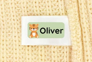 Namnlapp med en tiger som motiv och namnet Oliver, placerad på en mjuk, gyllene fleecebakgrund. Perfekt för att ge barnkläder eller tillbehör en personlig touch.