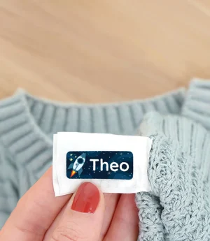 Navnelapp med navnet Theo i fokus, plassert på en blå strikkegenser med høstlige blader i bakgrunnen. Bilde viser hvordan navnelapper kan brukes til å identifisere klær for barn, et perfekt produkt for å unngå å miste eiendeler.